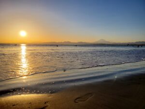 夕暮れの湘南海岸に輝く太陽と穏やかな波、奥には富士山のシルエットが映える風景